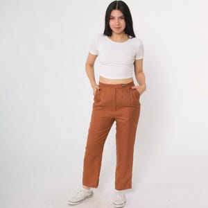 May include: A woman wearing a white short-sleeve crop top and rust-colored high-waisted pants. The pants have a button closure and pockets. She is wearing white sneakers. The background is plain white.
