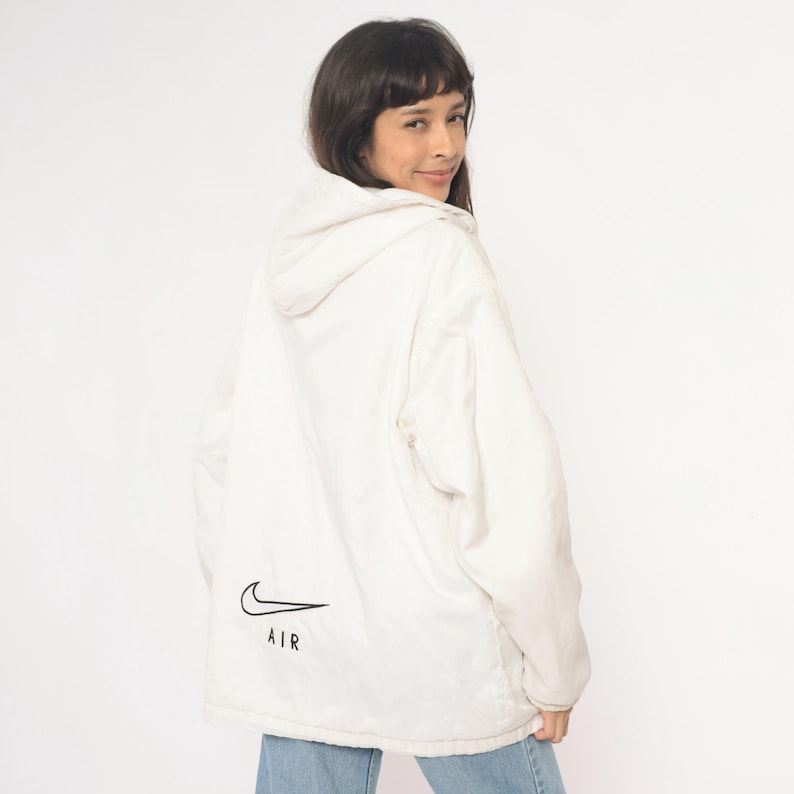 Puede incluir: Chaqueta con capucha blanca con el logo Nike Air negro en la espalda. La chaqueta la lleva una persona, sobre un fondo blanco liso. La chaqueta tiene una capucha con cord&oacute;n y un corte holgado.