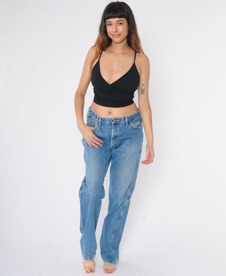 Pu&ograve; includere: Una donna indossa un top corto nero e jeans di denim blu chiaro. I jeans hanno una vestibilit&agrave; rilassata e una gamba dritta.