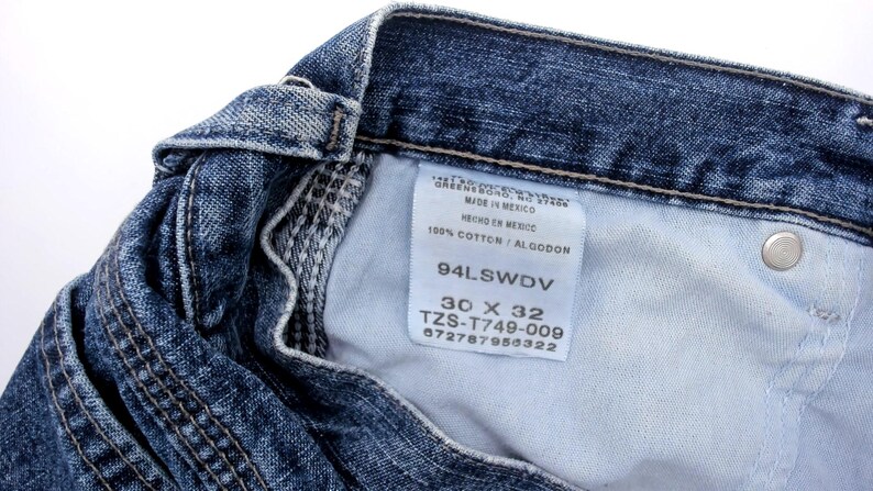 May include: Close-up of blue denim jeans with a white tag. The tag reads "MADE IN MEXICO" and "100% COTTON / ALGODON". The jeans are size 30 x 32.