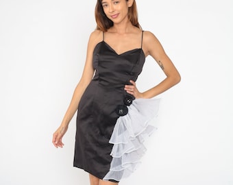May include: A black satin dress with a white tulle ruffle on the side. The dress has a sweetheart neckline and thin straps. There are two black fabric flowers on the tulle ruffle.