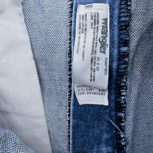 Puede incluir: Primer plano de unos vaqueros Wrangler de mezclilla azul. Los vaqueros tienen una etiqueta blanca con el logotipo de Wrangler y la informaci&oacute;n de la talla. La etiqueta dice "35 x 30". Los vaqueros est&aacute;n hechos en M&eacute;xico con tela estadounidense.