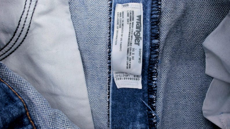 Puede incluir: Primer plano de unos vaqueros Wrangler de mezclilla azul. Los vaqueros tienen una etiqueta blanca con el logotipo de Wrangler y la informaci&oacute;n de la talla. La etiqueta dice "35 x 30". Los vaqueros est&aacute;n hechos en M&eacute;xico con tela estadounidense.