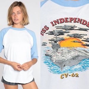 May include: White and pale blue raglan-sleeve t-shirt featuring the text "USS INDEPENDENCE CV-62" and an image of an aircraft carrier, sun, and clouds. The shirt has a crew neck and three-quarter length sleeves.