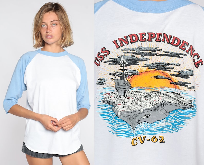 May include: White and pale blue raglan-sleeve t-shirt featuring the text "USS INDEPENDENCE CV-62" and an image of an aircraft carrier, sun, and clouds. The shirt has a crew neck and three-quarter length sleeves.