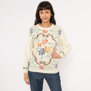 Peut inclure: Pull crème avec un motif floral. Le pull présente un motif floral central en rose, bleu et jaune, encadré d'une bordure marron. Les manches et l'encolure ont une texture côtelée. La personne porte un jean bleu.