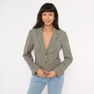 May include: A gray tweed blazer with two front pockets and two buttons. The blazer is worn with light blue jeans. The jacket has a classic, tailored design, suitable for various occasions.