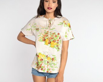 May include: A white blouse with a floral print featuring yellow, brown, and green flowers. The blouse has a tie at the neck and short sleeves.