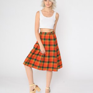 May include: A woman wearing a white tank top and a knee-length orange, green, and yellow plaid skirt. She is wearing beige platform sandals.