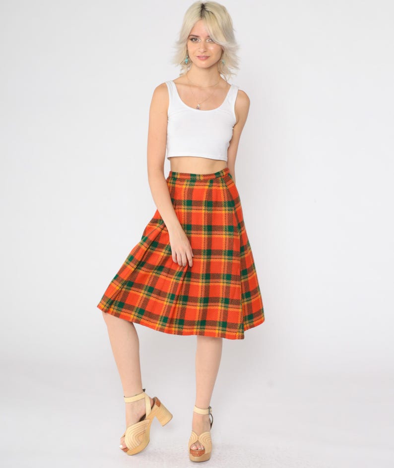 May include: A woman wearing a white tank top and a knee-length orange, green, and yellow plaid skirt. She is wearing beige platform sandals.