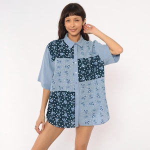 May include: A short-sleeve button-down shirt featuring a patchwork design of light blue and dark navy blue fabric panels. The shirt displays a repeating floral pattern across the different sections, a classic collar, and a front button closure.