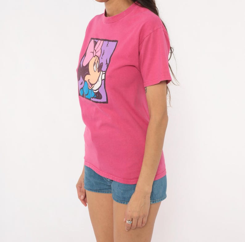 May include: A bright pink short-sleeved t-shirt featuring a rectangular graphic print of a cartoon character with a pink bow, wearing a blue dress, set against a purple background. The shirt is paired with blue denim shorts.