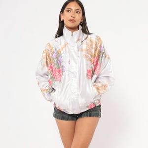 May include: A white vintage track jacket with a high collar and a full-length zipper. The jacket features a floral pattern in pink, purple, and gold. The jacket has pockets and elastic cuffs.