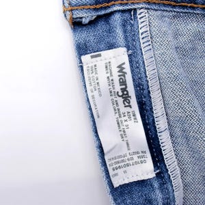 Puede incluir: Primer plano de unos vaqueros azules con una etiqueta blanca. La etiqueta dice "Wrangler" e incluye informaci&oacute;n sobre la talla: 34 x 31. Los vaqueros son 100% algod&oacute;n y se pueden lavar a m&aacute;quina. Es un estilo cl&aacute;sico.