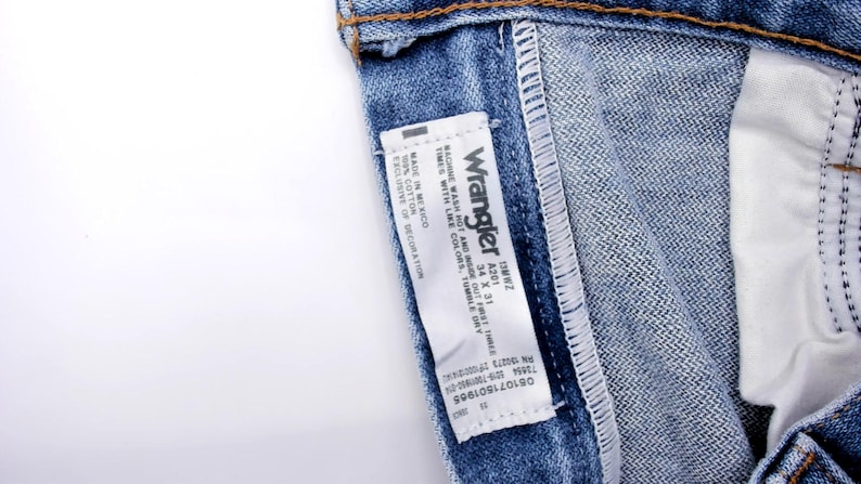 Puede incluir: Primer plano de unos vaqueros azules con una etiqueta blanca. La etiqueta dice "Wrangler" e incluye informaci&oacute;n sobre la talla: 34 x 31. Los vaqueros son 100% algod&oacute;n y se pueden lavar a m&aacute;quina. Es un estilo cl&aacute;sico.