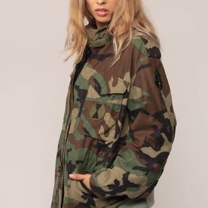 May include: A camouflage field jacket featuring a woodland pattern in shades of green, brown, and black. The jacket includes a high collar, a front zipper closure, two chest pockets with flaps, and circular patches on the chest and sleeve.