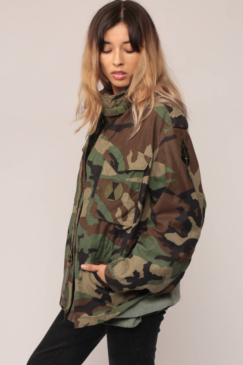 May include: A camouflage field jacket featuring a woodland pattern in shades of green, brown, and black. The jacket includes a high collar, a front zipper closure, two chest pockets with flaps, and circular patches on the chest and sleeve.