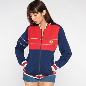 Peut inclure: Une veste bomber rayée bleu et rouge avec une fermeture éclair blanche et un petit logo sur la poitrine. La veste a des bordures blanches autour du col, des poignets et des poches.