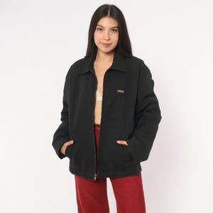 May include: A black zip-up jacket with a pointed collar, long sleeves, and two front pockets. The jacket features a small rectangular brand label on the left chest area. The garment is worn open over a light-colored top and red pants.