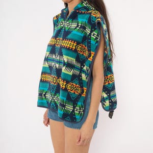 May include: A sleeveless hooded poncho featuring a vibrant geometric pattern in shades of navy blue, teal, bright green, yellow, and orange. The garment has an open-side design with dark blue trim along the edges and a hood at the neckline.