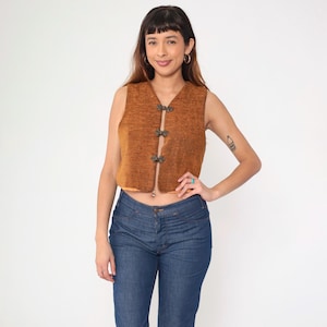 May include: A sleeveless, brown vest with decorative clasps. The vest is made of a textured fabric and has a V-neck. The vest is worn with blue jeans.