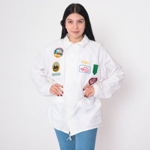 Op de afbeelding: Witte jas met verschillende kleurrijke patches, waaronder een patch met de tekst "Debbie" en een andere patch met de tekst "Ashley I Love You".