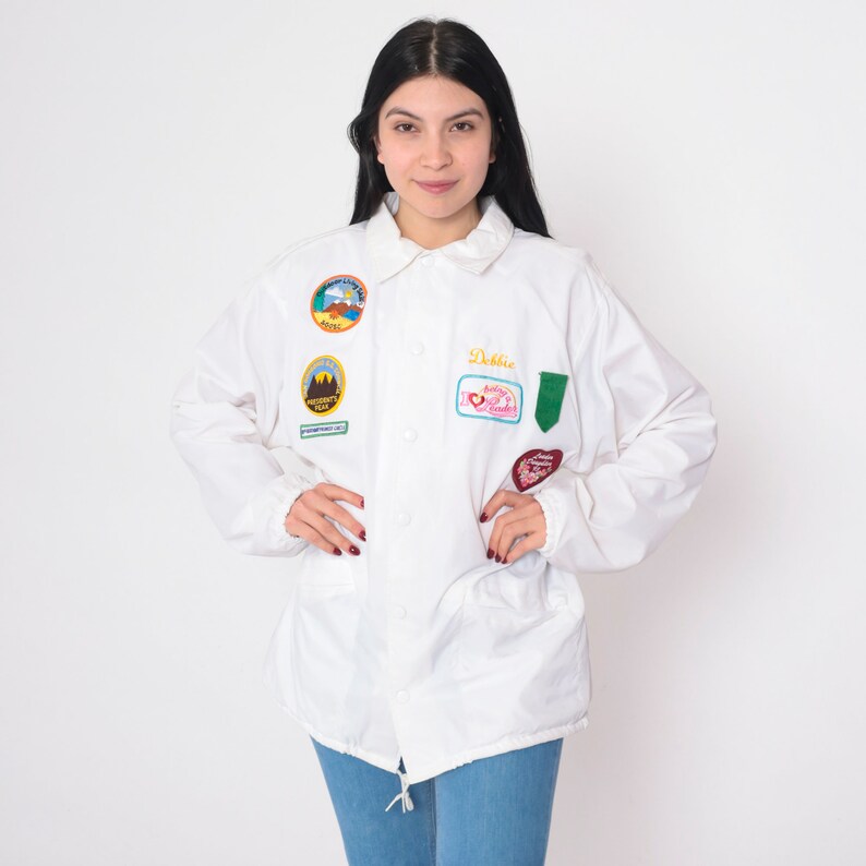 Op de afbeelding: Witte jas met verschillende kleurrijke patches, waaronder een patch met de tekst "Debbie" en een andere patch met de tekst "Ashley I Love You".
