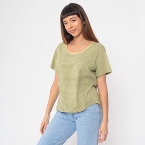 Peut inclure: Un t-shirt &agrave; rayures vertes et blanches avec un col rond et des manches courtes. Le t-shirt a une bordure jaune autour de l'encolure et est associ&eacute; &agrave; un jean bleu clair. Le mod&egrave;le sourit.