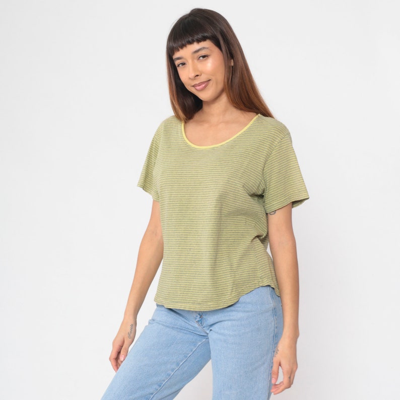 Peut inclure: Un t-shirt &agrave; rayures vertes et blanches avec un col rond et des manches courtes. Le t-shirt a une bordure jaune autour de l'encolure et est associ&eacute; &agrave; un jean bleu clair. Le mod&egrave;le sourit.