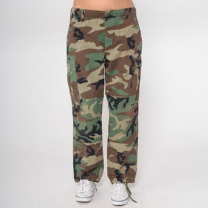 May include: Camouflage cargo pants in shades of green, brown, and black. The pants have multiple pockets and a straight leg fit. The model is wearing white sneakers. The pants are made of a durable fabric.