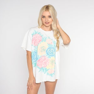 Puede incluir: Una camiseta blanca de gran tama&ntilde;o con un estampado floral en rosa pastel, azul y amarillo. Las flores son estilizadas y tienen un aspecto pict&oacute;rico.