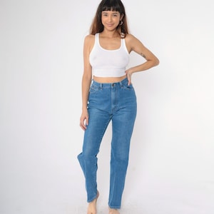 Op de afbeelding: Een vrouw draagt een wit tanktop en een blauwe spijkerbroek. De spijkerbroek heeft een hoge taille en een rechte pijp.