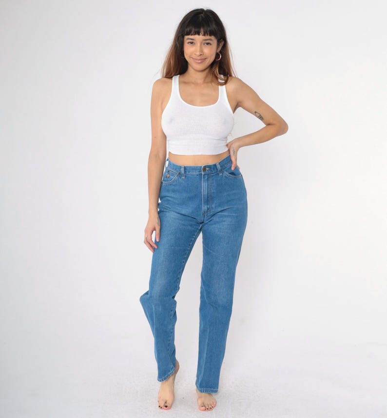 Op de afbeelding: Een vrouw draagt een wit tanktop en een blauwe spijkerbroek. De spijkerbroek heeft een hoge taille en een rechte pijp.