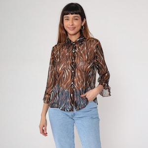 May include: A sheer button-down shirt with a brown and black zebra print. The shirt has a collared neckline, long sleeves, and is paired with light blue jeans. The model is wearing gold hoop earrings.