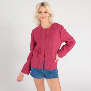 May include: A magenta-colored jacket with a scalloped design down the front, fastened with six buttons. The jacket has long sleeves and a round neckline. The model is wearing blue denim shorts.