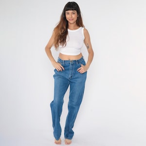 May include: A woman wearing a white tank top and blue denim jeans. The jeans have a high waist and a straight leg.