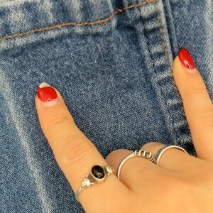 May include: Close-up of blue denim fabric with a visible seam and stitching. A hand with red nail polish and silver rings is pointing towards the denim. The rings include a black stone and geometric designs.