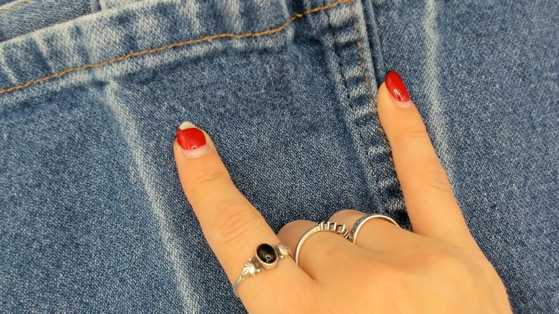 May include: Close-up of blue denim fabric with a visible seam and stitching. A hand with red nail polish and silver rings is pointing towards the denim. The rings include a black stone and geometric designs.
