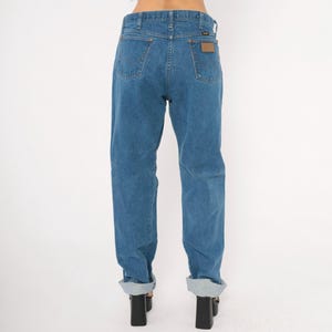 May include: Blue denim jeans with a classic fit, featuring a brown leather patch on the back pocket. The jeans have a straight leg and are cuffed at the ankles. The jeans are a medium wash denim.