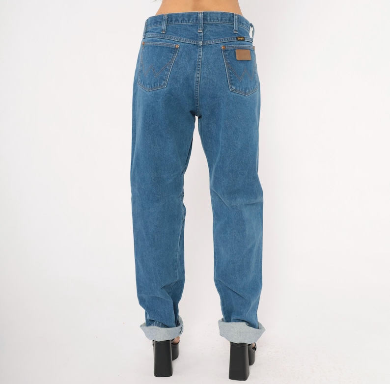 May include: Blue denim jeans with a classic fit, featuring a brown leather patch on the back pocket. The jeans have a straight leg and are cuffed at the ankles. The jeans are a medium wash denim.
