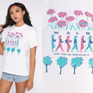 May include: White t-shirt featuring a graphic of people walking and flowers. The text "WALK JUST FOR THE HEALTH OF IT" is printed on the shirt.
