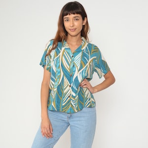 May include: Short-sleeved button-up shirt with a teal, white, and gold leaf pattern. The shirt has a collared neckline and wooden buttons. The model is wearing light blue jeans.