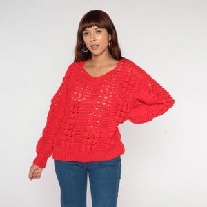 May include: A bright red, open-weave sweater with a wide, scoop neckline. The long-sleeved top has a loose, relaxed fit. The model is wearing blue jeans. The sweater is a vibrant, eye-catching piece.