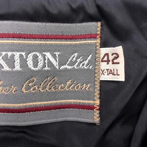 May include: Close-up of a black leather garment tag. The tag is gray with the words "OAKTON Ltd. Leather Collection" embroidered in white and tan. A smaller tag reads "42 X-TALL".