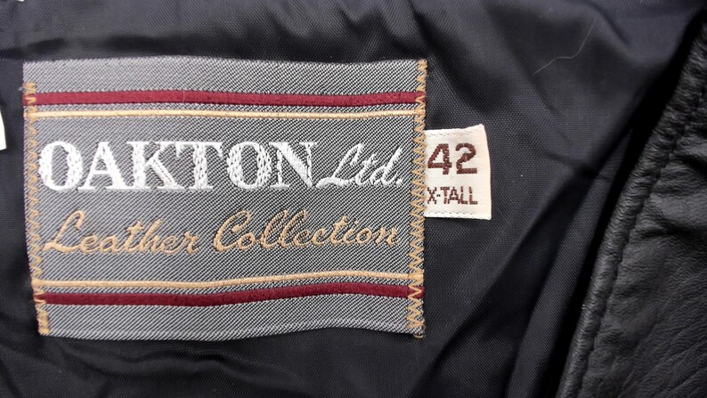 May include: Close-up of a black leather garment tag. The tag is gray with the words "OAKTON Ltd. Leather Collection" embroidered in white and tan. A smaller tag reads "42 X-TALL".