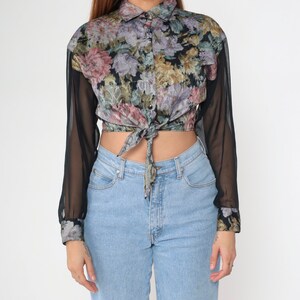 May include: A floral print button-up shirt with sheer black sleeves tied at the waist. The shirt features a colorful floral pattern with pink, purple, and yellow flowers. The sleeves are long and sheer, with floral cuffs. The shirt is paired with light blue jeans.