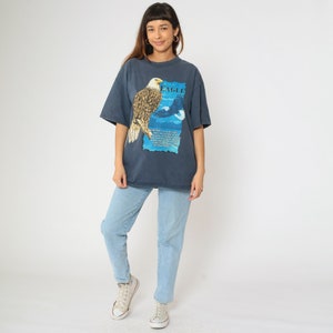 May include: A dark blue t-shirt featuring a graphic print of a bald eagle with the text "Bald Eagle" on the front. The eagle is perched on a branch with its wings spread. The background is a blue sky with clouds.