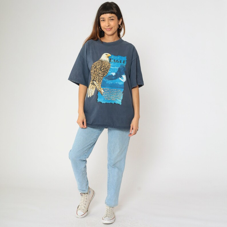 May include: A dark blue t-shirt featuring a graphic print of a bald eagle with the text "Bald Eagle" on the front. The eagle is perched on a branch with its wings spread. The background is a blue sky with clouds.