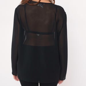 May include: Black sheer long-sleeve top. The top has a loose fit and a scoop neckline. The fabric is lightweight and see-through. The sleeves are long and end at the wrists. The top is worn over a black bra and black pants.