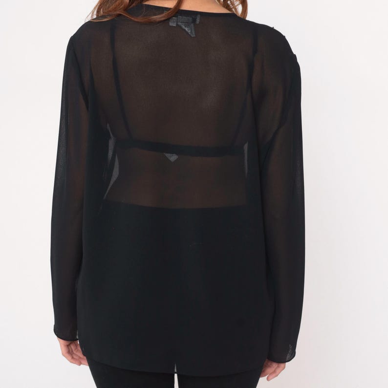 May include: Black sheer long-sleeve top. The top has a loose fit and a scoop neckline. The fabric is lightweight and see-through. The sleeves are long and end at the wrists. The top is worn over a black bra and black pants.